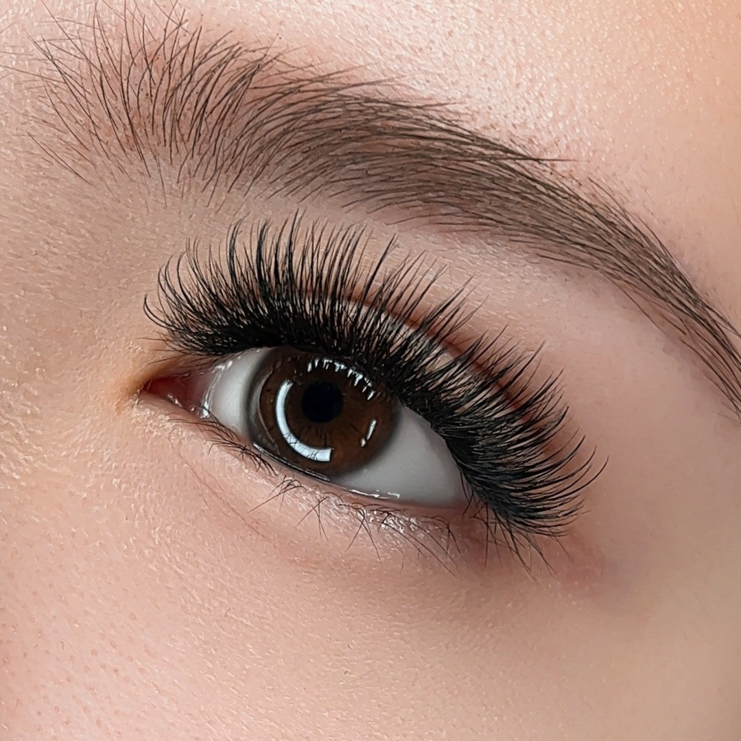 Close-up of a human eye wearing dramatic, long, 7D Wispy Promade lash extensions with a neatly shaped eyebrow and smooth skin.