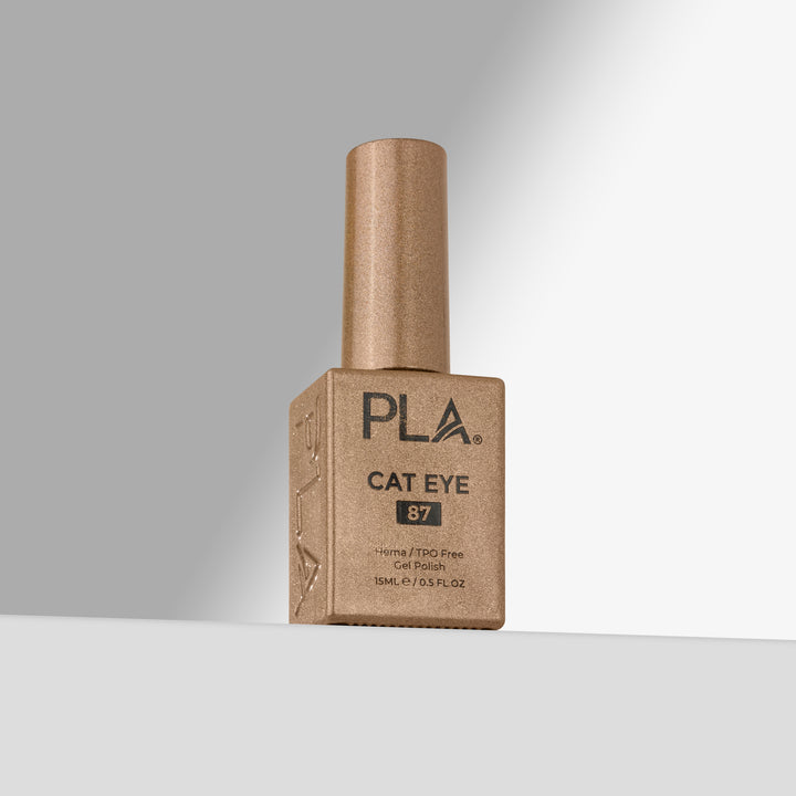 A bottle of PLA Cat Eye 87 nail polish in a gold shade, standing upright on a light gray surface against a gray background.