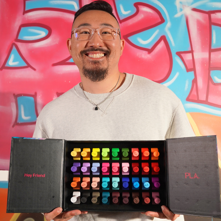 A smiling man (Shawn) wearing glasses and a light gray sweatshirt holds open a black box of colorful craft sticks or paint-like markers labeled &